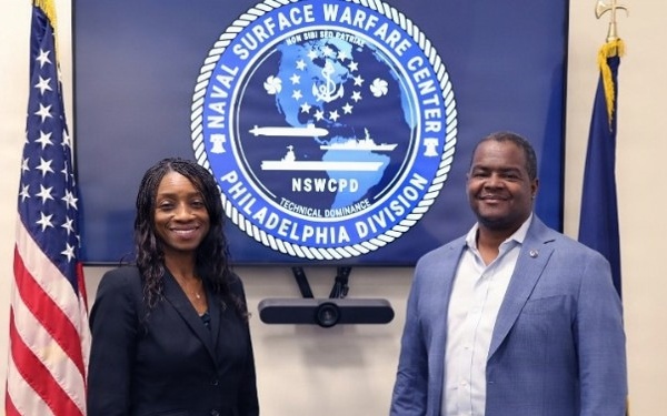 Two individuals are posing for a photo in front of a large display screen showing the official seal of the Naval Surface Warfare Center, Philadelphia Division (NSWCPD). The setting appears to be an official meeting room or conference space within the facility. An American flag and a Navy flag are visible flanking the display.