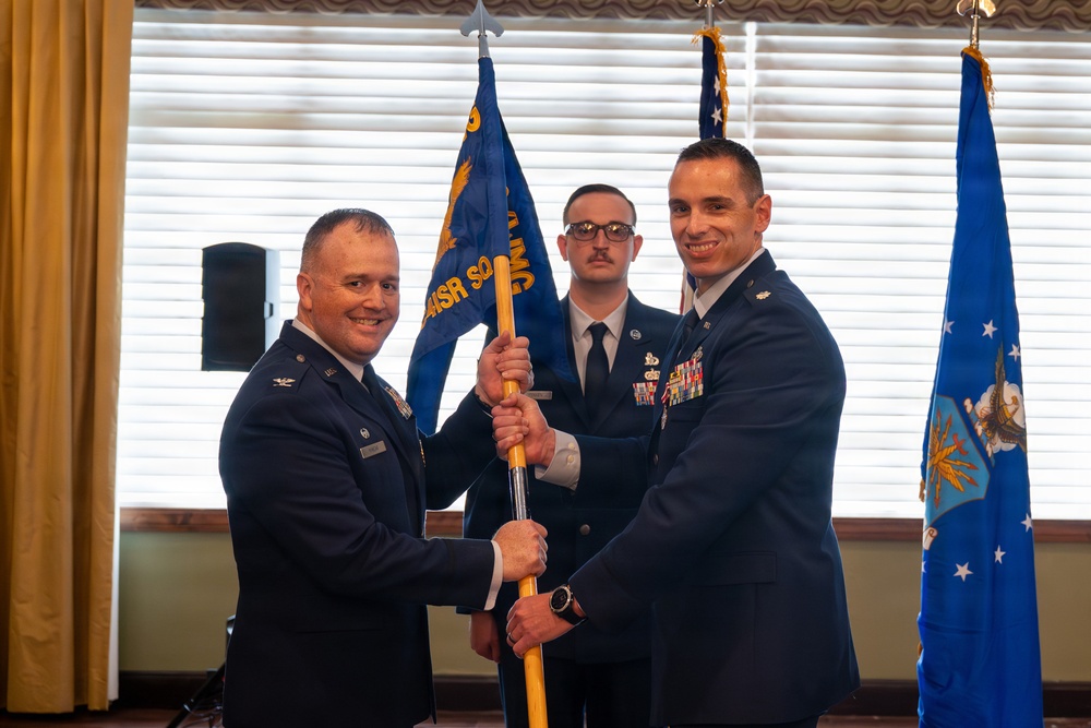 Two U.S. Air Force officers are participating in a formal guidon-passing ceremony, likely a change of command. A third uniformed airman stands in the background holding the unit guidon staff. The setting appears to be an indoor venue with U.S. Air Force flags displayed.