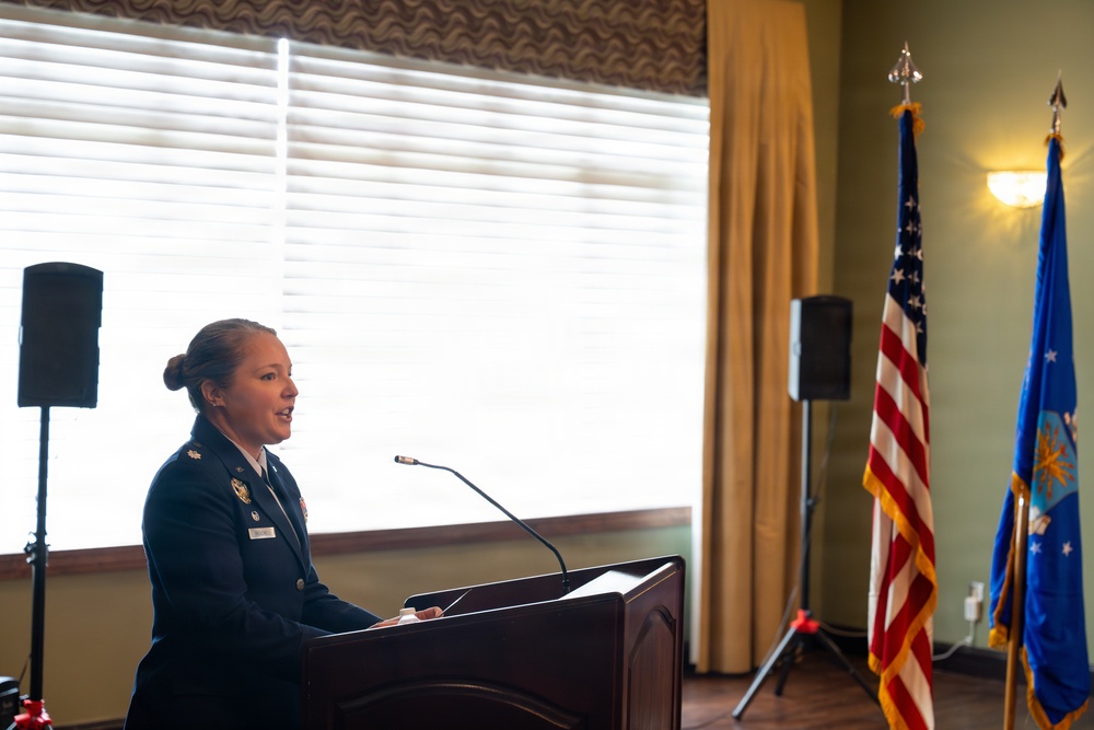 A female military officer in dress uniform is speaking at a podium during what appears to be an official ceremony or formal event. She is positioned in front of a large window with blinds, flanked by the United States flag and what appears to be the United States Air Force flag. The setting suggests an indoor military function or awards ceremony.