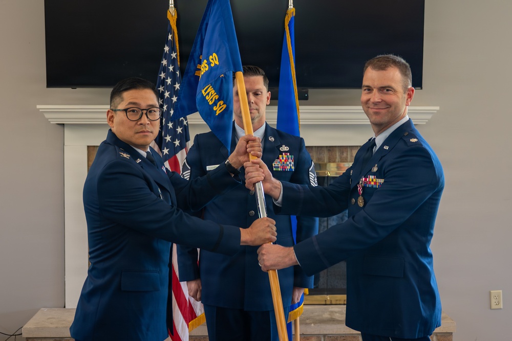 Three U.S. Air Force personnel in service dress uniforms are participating in a formal change of command or activation ceremony. Two officers in the foreground are jointly holding a unit guidon (flag on a staff), while a senior non-commissioned officer stands in the background holding the guidon pole. The ceremony is taking place indoors in front of an American flag and a blue Air Force unit flag.