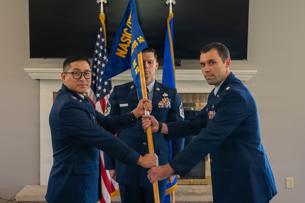 A military change of command ceremony is taking place indoors, with two Air Force officers transferring a unit guidon (flag). The blue guidon bears the text 'NASIC' and 'SQ', indicating a National Air and Space Intelligence Center squadron. A third service member stands at attention in the background holding additional flags.