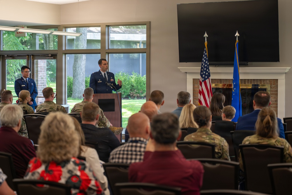 A U.S. Air Force officer in dress blues is delivering a speech at a podium during what appears to be an official military ceremony or change of command event. An audience of mixed military personnel in various uniforms and civilians is seated facing the speaker. Two flags — the U.S. national flag and a U.S. Air Force flag — are displayed prominently beside a fireplace and large wall-mounted television screen.