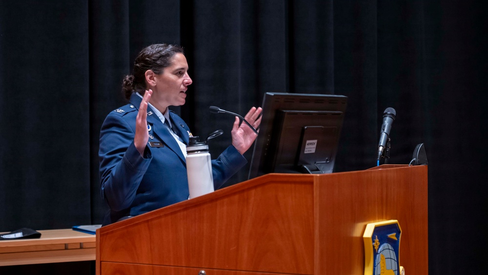 A female military officer in a blue dress uniform is delivering a presentation or speech at a wooden podium. She is gesturing with both hands raised, suggesting active engagement with her audience. A military insignia is visible on the front of the podium.