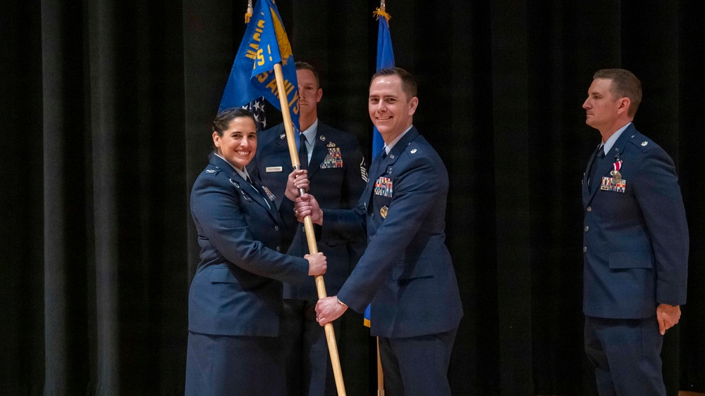 A military change of command ceremony is taking place on a stage, with two officers jointly holding a unit guidon (flag). The central action shows the formal passing of the guidon between a female officer and a male officer, a traditional symbol of transferring command authority. Additional uniformed personnel stand in the background observing the ceremony.