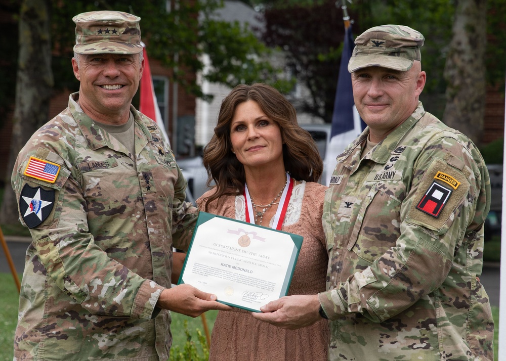 Two U.S. Army soldiers in OCP (Operational Camouflage Pattern) uniforms are presenting a Department of the Army certificate to a civilian woman named Katie McDonald. The certificate appears to be a Meritorious Public Service Medal award. The ceremony is taking place outdoors with flags visible in the background.