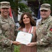 Two U.S. Army soldiers in OCP (Operational Camouflage Pattern) uniforms are presenting a Department of the Army certificate to a civilian woman named Katie McDonald. The certificate appears to be a Meritorious Public Service Medal award. The ceremony is taking place outdoors with flags visible in the background.