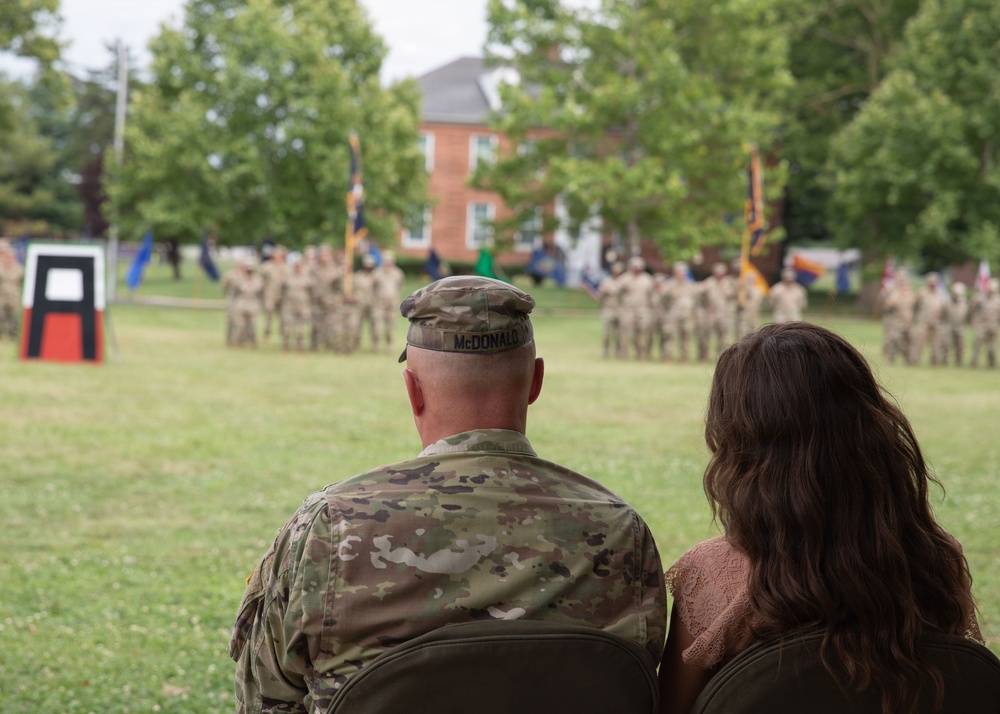 A U.S. Army soldier named McDonald, wearing OCP (Operational Camouflage Pattern) uniform, sits alongside a civilian woman observing a military formation ceremony on a parade field. In the background, a large formation of soldiers in uniform stands at attention with multiple unit guidons and flags visible. A unit insignia display board is visible on the left side of the field.