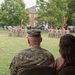 A U.S. Army soldier named McDonald, wearing OCP (Operational Camouflage Pattern) uniform, sits alongside a civilian woman observing a military formation ceremony on a parade field. In the background, a large formation of soldiers in uniform stands at attention with multiple unit guidons and flags visible. A unit insignia display board is visible on the left side of the field.