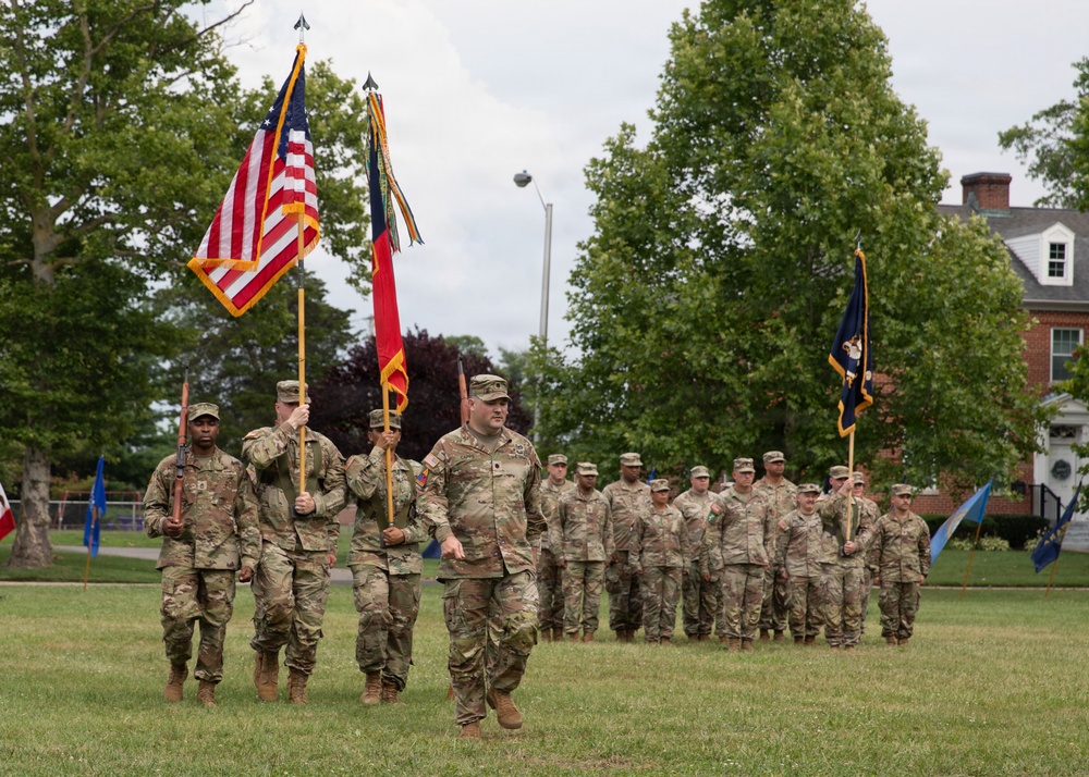 A military color guard and formation are conducting a formal ceremony on a grass parade field. Soldiers in OCP (Operational Camouflage Pattern) uniforms are marching with multiple flags including the U.S. national flag, a red unit guidon, and a blue flag. The setting appears to be a military installation with brick buildings and mature trees visible in the background.