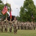 A military color guard and formation are conducting a formal ceremony on a grass parade field. Soldiers in OCP (Operational Camouflage Pattern) uniforms are marching with multiple flags including the U.S. national flag, a red unit guidon, and a blue flag. The setting appears to be a military installation with brick buildings and mature trees visible in the background.