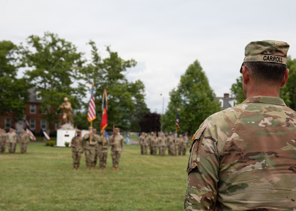 A military ceremony is taking place on a grassy parade field, with a soldier named Carroll visible in the foreground observing the proceedings. Multiple soldiers in OCP (Operational Camouflage Pattern) uniforms are assembled in formation in the background, with color guard bearers holding flags including the American flag. A gold/bronze military statue is visible on a pedestal in the background, suggesting this may be a formal installation or unit ceremony.