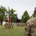 A military ceremony is taking place on a grassy parade field, with a soldier named Carroll visible in the foreground observing the proceedings. Multiple soldiers in OCP (Operational Camouflage Pattern) uniforms are assembled in formation in the background, with color guard bearers holding flags including the American flag. A gold/bronze military statue is visible on a pedestal in the background, suggesting this may be a formal installation or unit ceremony.