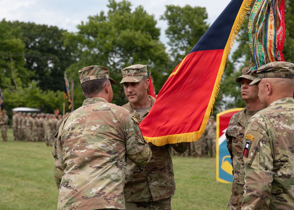 A U.S. Army change of command or activation ceremony is taking place on a grassy parade field. Soldiers in OCP (Operational Camouflage Pattern) uniforms are passing a unit colors/guidon flag with gold fringe, which is the central ritual of a change of command ceremony. Additional soldiers and unit flags are visible in formation in the background.