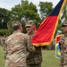 A U.S. Army change of command or activation ceremony is taking place on a grassy parade field. Soldiers in OCP (Operational Camouflage Pattern) uniforms are passing a unit colors/guidon flag with gold fringe, which is the central ritual of a change of command ceremony. Additional soldiers and unit flags are visible in formation in the background.