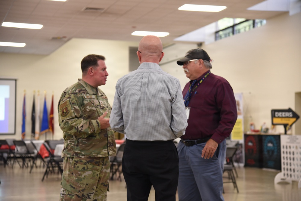 Three men are engaged in conversation in what appears to be a military community event or resource fair. One man is wearing a U.S. military camouflage uniform (OCP pattern), while the other two are in civilian attire. The setting includes military branch flags, folding chairs, and informational booths in the background.