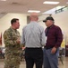 Three men are engaged in conversation in what appears to be a military community event or resource fair. One man is wearing a U.S. military camouflage uniform (OCP pattern), while the other two are in civilian attire. The setting includes military branch flags, folding chairs, and informational booths in the background.