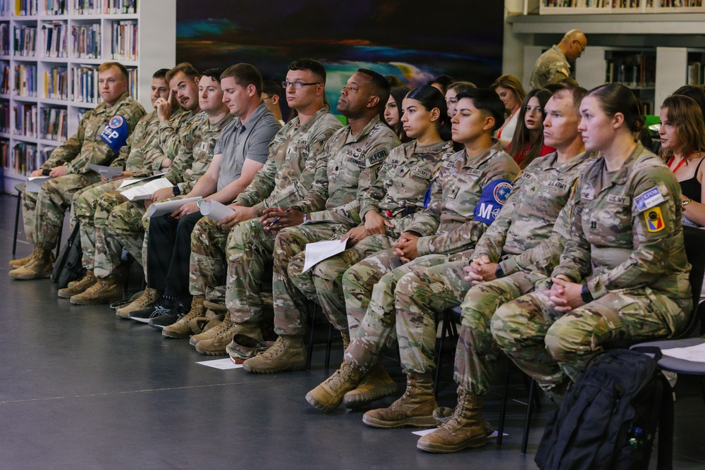 A group of military personnel in camouflage uniforms (OCP/MultiCam pattern) are seated in rows in what appears to be a library or educational facility. The majority are U.S. Army soldiers, with at least one Air Force member visible, along with a few civilians seated behind them. They appear to be attending a briefing, presentation, or formal event.