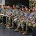 A group of military personnel in camouflage uniforms (OCP/MultiCam pattern) are seated in rows in what appears to be a library or educational facility. The majority are U.S. Army soldiers, with at least one Air Force member visible, along with a few civilians seated behind them. They appear to be attending a briefing, presentation, or formal event.
