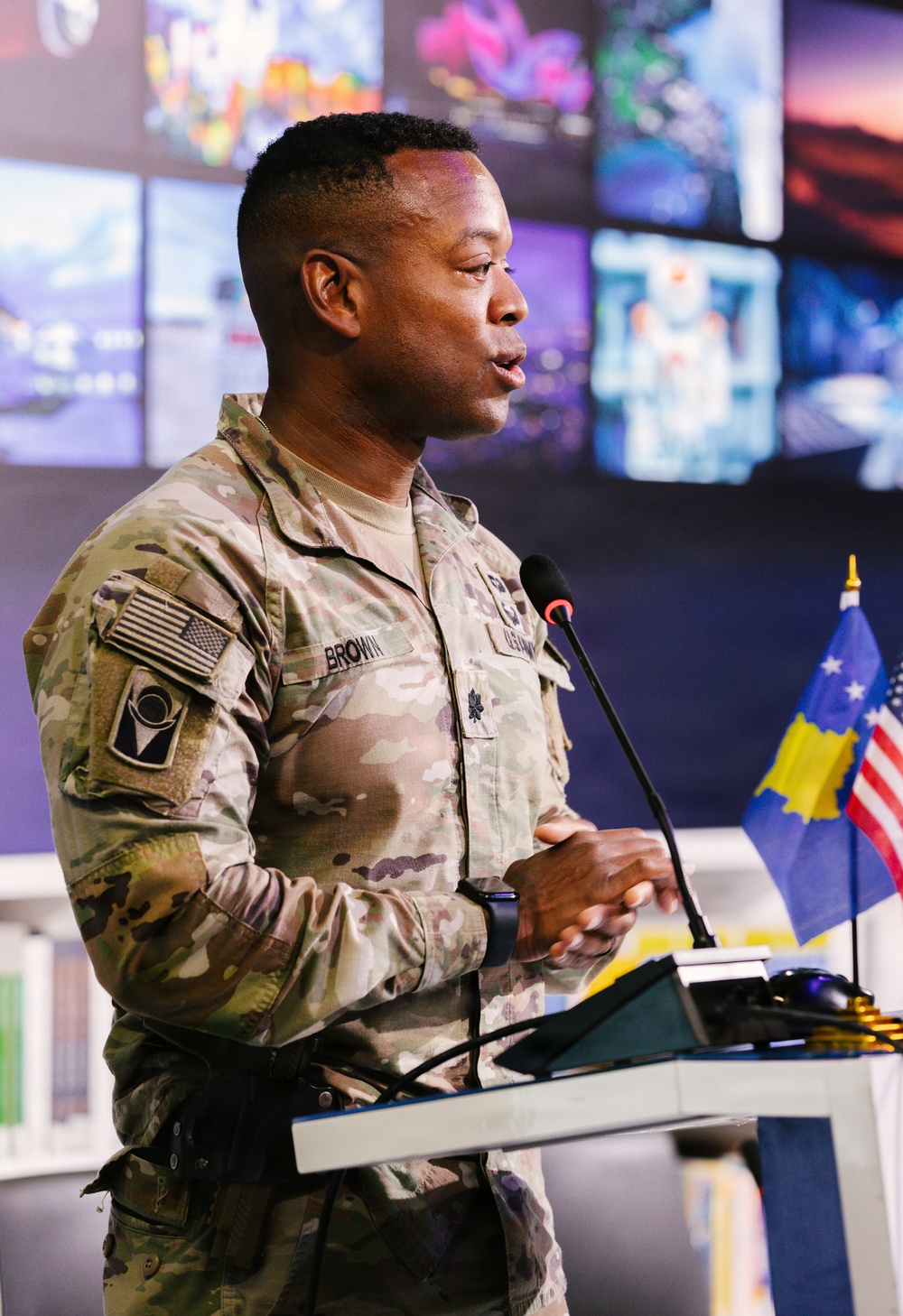 A U.S. Army officer with the name tape 'BROWN' and rank insignia of Lieutenant Colonel (oak leaf cluster) is speaking at a podium equipped with a microphone. He is wearing the Army Combat Uniform (ACU) in MultiCam pattern with a U.S. flag patch on the right shoulder. Behind him are small flags including the Kosovo flag and the U.S. flag, along with a wall of multiple display screens.
