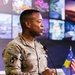 A U.S. Army officer with the name tape 'BROWN' and rank insignia of Lieutenant Colonel (oak leaf cluster) is speaking at a podium equipped with a microphone. He is wearing the Army Combat Uniform (ACU) in MultiCam pattern with a U.S. flag patch on the right shoulder. Behind him are small flags including the Kosovo flag and the U.S. flag, along with a wall of multiple display screens.