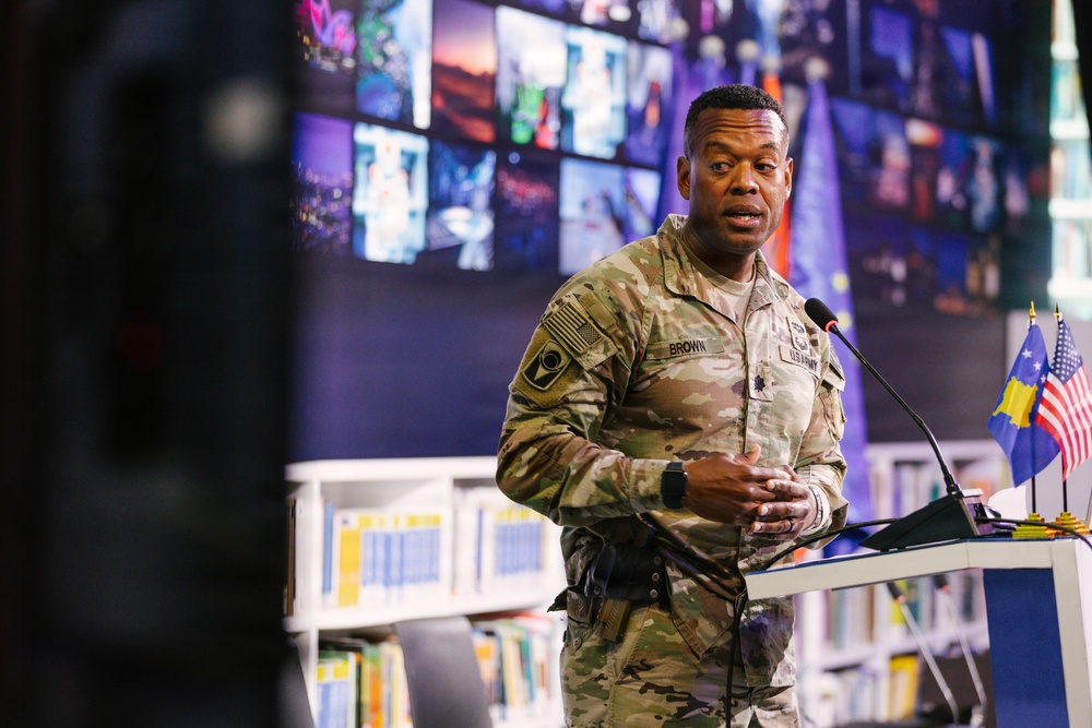 A U.S. Army soldier named Brown is speaking at a podium in what appears to be a library or media center. Two small flags — one American and one appearing to be the flag of Kosovo — are visible on the podium. The setting suggests an official military engagement or community outreach event.