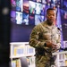 A U.S. Army soldier named Brown is speaking at a podium in what appears to be a library or media center. Two small flags — one American and one appearing to be the flag of Kosovo — are visible on the podium. The setting suggests an official military engagement or community outreach event.