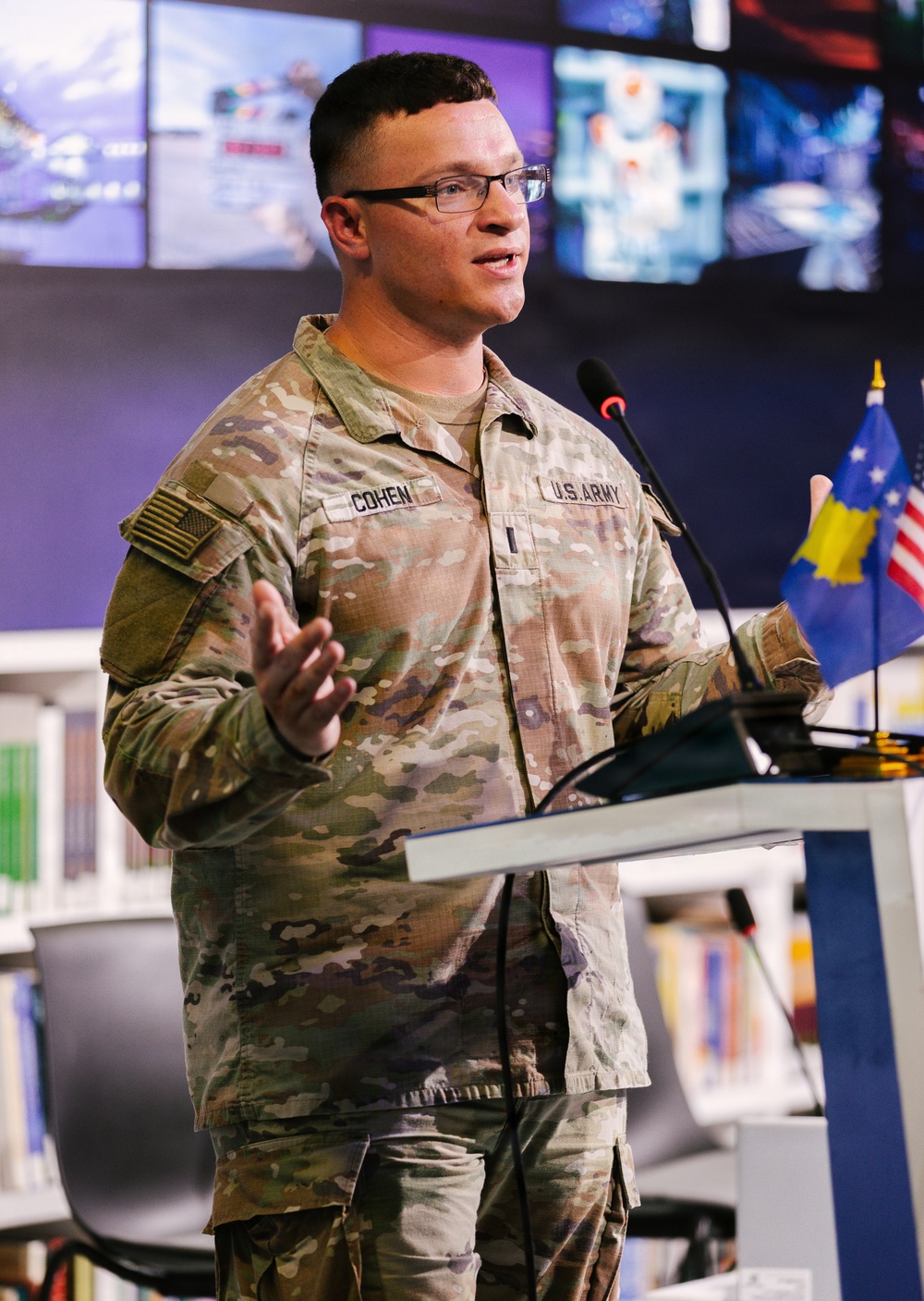 A U.S. Army soldier named Cohen is speaking at a podium in what appears to be a modern briefing room or library setting. He is wearing MultiCam (OCP) camouflage uniform with U.S. Army and name tape visible, along with a single rank insignia pin. Two small flags — one appearing to be the Kosovo flag and one the U.S. flag — are visible on the podium.
