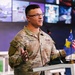 A U.S. Army soldier named Cohen is speaking at a podium in what appears to be a modern briefing room or library setting. He is wearing MultiCam (OCP) camouflage uniform with U.S. Army and name tape visible, along with a single rank insignia pin. Two small flags — one appearing to be the Kosovo flag and one the U.S. flag — are visible on the podium.
