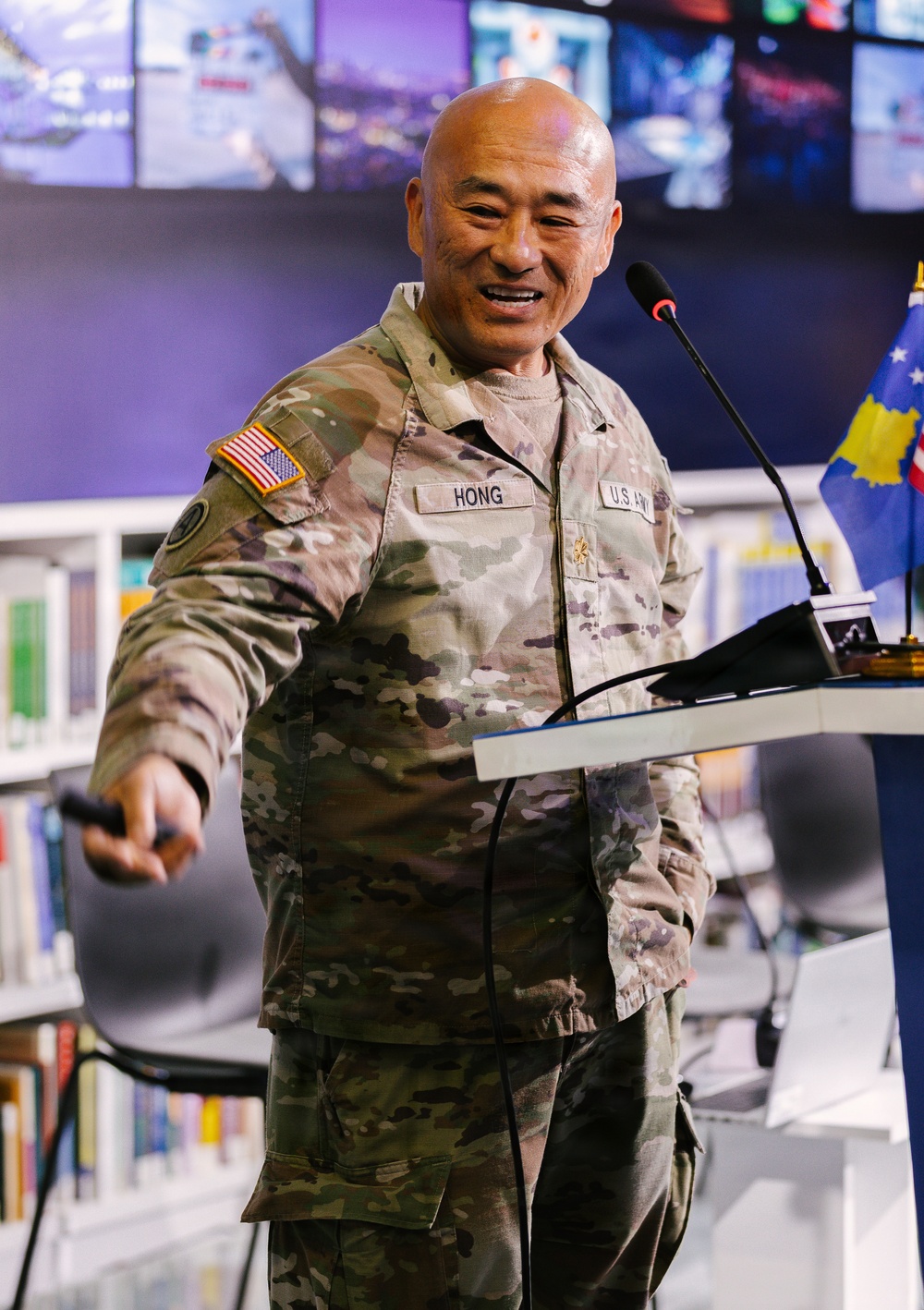 A U.S. Army officer named HONG, wearing OCP (Operational Camouflage Pattern) uniform, is speaking at a podium with a microphone. He is smiling and gesturing with his right hand while holding what appears to be a presentation clicker. The setting appears to be a modern briefing room or library with a video wall display in the background.
