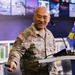 A U.S. Army officer named HONG, wearing OCP (Operational Camouflage Pattern) uniform, is speaking at a podium with a microphone. He is smiling and gesturing with his right hand while holding what appears to be a presentation clicker. The setting appears to be a modern briefing room or library with a video wall display in the background.