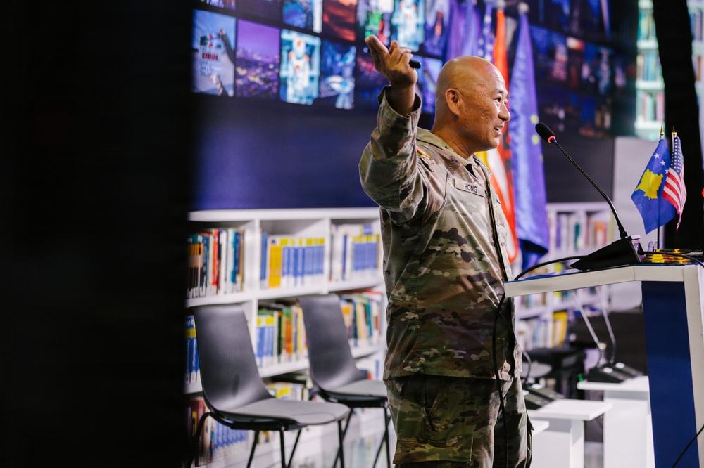 A U.S. Army soldier with the name tape 'HONG' is delivering a presentation or speech at a podium in what appears to be a library or educational facility. He is wearing OCP (Operational Camouflage Pattern) uniform and gesturing with his right hand while holding a presentation clicker. Two small flags — one American and one Kosovo — are visible on the podium.