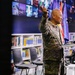 A U.S. Army soldier with the name tape 'HONG' is delivering a presentation or speech at a podium in what appears to be a library or educational facility. He is wearing OCP (Operational Camouflage Pattern) uniform and gesturing with his right hand while holding a presentation clicker. Two small flags — one American and one Kosovo — are visible on the podium.