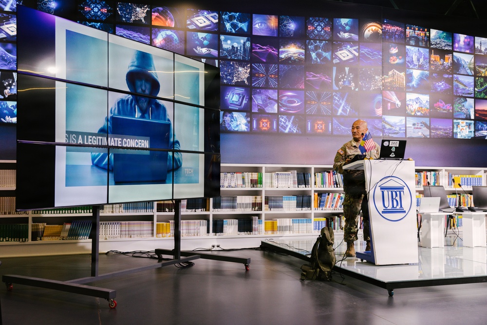 A military personnel in camouflage uniform is delivering a presentation at a podium marked 'UBT' in what appears to be a modern library or media center. The large display screen behind them shows an image of a hooded figure at a laptop with the text 'IS A LEGITIMATE CONCERN', suggesting a cybersecurity-themed lecture. The backdrop features an extensive video wall displaying various technology-related images.