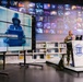 A military personnel in camouflage uniform is delivering a presentation at a podium marked 'UBT' in what appears to be a modern library or media center. The large display screen behind them shows an image of a hooded figure at a laptop with the text 'IS A LEGITIMATE CONCERN', suggesting a cybersecurity-themed lecture. The backdrop features an extensive video wall displaying various technology-related images.