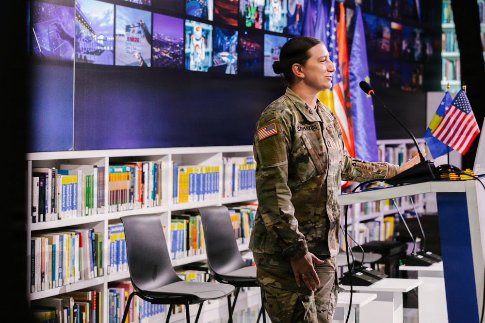 A female U.S. Army soldier in MultiCam/OCP camouflage uniform is speaking at a podium in what appears to be a modern library or educational facility. Small U.S. and Kosovo flags are visible on the podium, and large international flags are displayed in the background. The setting suggests an official military or diplomatic engagement event.