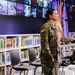 A female U.S. Army soldier in MultiCam/OCP camouflage uniform is speaking at a podium in what appears to be a modern library or educational facility. Small U.S. and Kosovo flags are visible on the podium, and large international flags are displayed in the background. The setting suggests an official military or diplomatic engagement event.