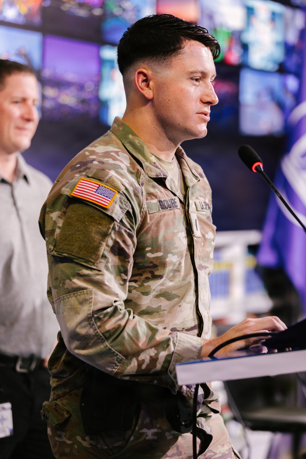 A U.S. Army soldier in Operational Camouflage Pattern (OCP) uniform is standing at a podium with a microphone, appearing to speak or present. The soldier has a U.S. flag patch on the right sleeve and a name tape visible on the chest. The setting appears to be a modern operations or media center with multiple display screens visible in the background.