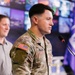 A U.S. Army soldier in Operational Camouflage Pattern (OCP) uniform is standing at a podium with a microphone, appearing to speak or present. The soldier has a U.S. flag patch on the right sleeve and a name tape visible on the chest. The setting appears to be a modern operations or media center with multiple display screens visible in the background.