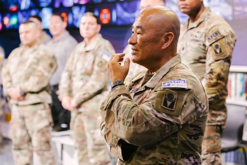 A group of military personnel in U.S. Army MultiCam/OCP camouflage uniforms are gathered in what appears to be an operations or briefing room. The central figure, a bald male soldier, is in a contemplative pose with his hand raised to his chin, appearing to listen or think. Patches visible on uniforms indicate KFOR (Kosovo Force) and RC-East (Regional Command East) affiliation.