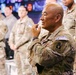 A group of military personnel in U.S. Army MultiCam/OCP camouflage uniforms are gathered in what appears to be an operations or briefing room. The central figure, a bald male soldier, is in a contemplative pose with his hand raised to his chin, appearing to listen or think. Patches visible on uniforms indicate KFOR (Kosovo Force) and RC-East (Regional Command East) affiliation.
