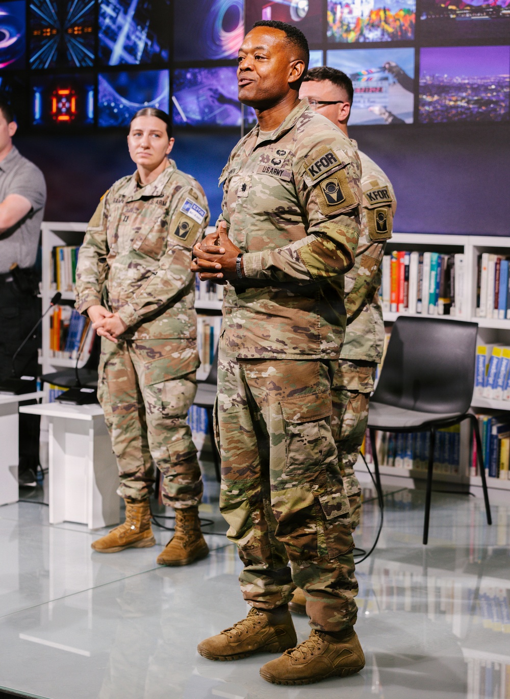 Three U.S. Army soldiers in OCP (Operational Camouflage Pattern) uniforms are standing in what appears to be a modern media studio or library setting. The central figure, a senior officer identifiable by rank insignia and 'KFOR' patch, appears to be speaking or presenting. The setting includes bookshelves and a large video display wall in the background.