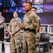 Three U.S. Army soldiers in OCP (Operational Camouflage Pattern) uniforms are standing in what appears to be a modern media studio or library setting. The central figure, a senior officer identifiable by rank insignia and 'KFOR' patch, appears to be speaking or presenting. The setting includes bookshelves and a large video display wall in the background.