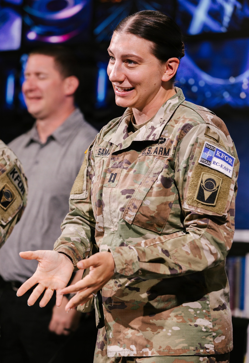 A U.S. Army officer in OCP (Operational Camouflage Pattern) uniform is smiling and gesturing with open hands, appearing to be speaking or presenting. She wears NATO KFOR RC-East patches on her right shoulder, indicating deployment to Kosovo Force. A civilian male in a gray shirt is visible in the background.
