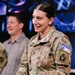 A U.S. Army officer in OCP (Operational Camouflage Pattern) uniform is smiling and gesturing with open hands, appearing to be speaking or presenting. She wears NATO KFOR RC-East patches on her right shoulder, indicating deployment to Kosovo Force. A civilian male in a gray shirt is visible in the background.