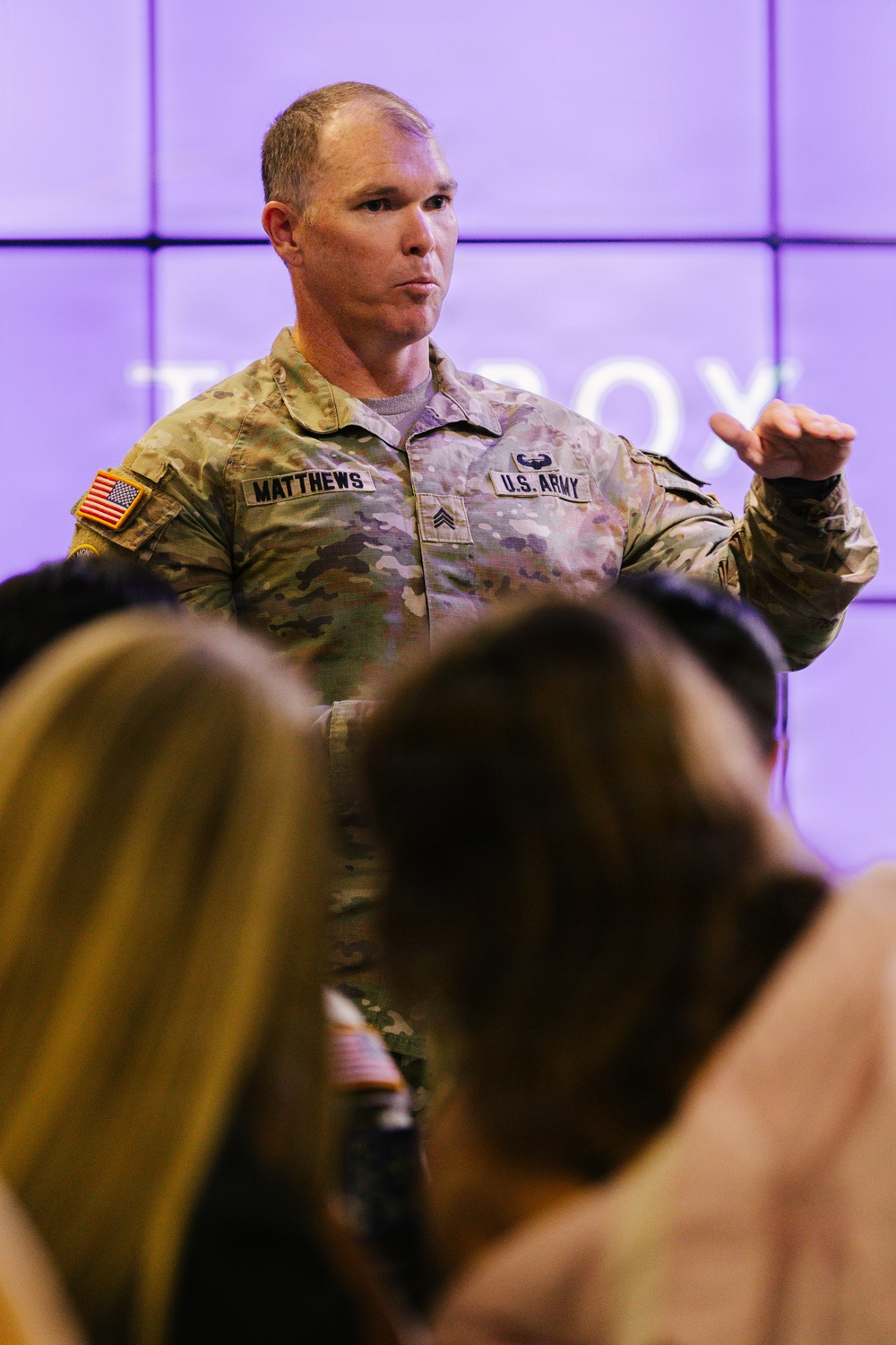 A U.S. Army Sergeant named Matthews is addressing an audience in what appears to be a presentation or briefing setting. He is wearing the Army Combat Uniform (ACU) in OCP (Operational Camouflage Pattern) with visible name tape, branch tape, U.S. flag patch, and rank insignia. Several audience members are visible in the foreground, out of focus.