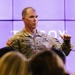 A U.S. Army Sergeant named Matthews is addressing an audience in what appears to be a presentation or briefing setting. He is wearing the Army Combat Uniform (ACU) in OCP (Operational Camouflage Pattern) with visible name tape, branch tape, U.S. flag patch, and rank insignia. Several audience members are visible in the foreground, out of focus.