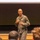 A bald male military officer wearing an olive/sage green flight suit is addressing an audience in what appears to be a lecture hall or briefing room. He is gesturing with both hands while speaking, standing in front of a large projection screen and chalkboard. Several audience members are visible from behind in the foreground.