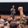A military officer in a flight suit is presenting or speaking to an audience of uniformed military personnel in a lecture hall or auditorium. One audience member in the foreground has raised a fist, likely to ask a question or respond to the speaker. The speaker appears to be pointing toward the audience, possibly acknowledging the raised hand.