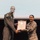 Two military personnel are posing together on a stage while jointly holding a framed award certificate. The individual on the left is wearing an olive/tan flight suit with patches, while the individual on the right is wearing a camouflage uniform with a name tag reading 'PARDO'. The framed certificate appears to read 'Amelia Earhart Award' at the top.