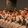 A group of young people wearing military-style camouflage uniforms, identifiable as Civil Air Patrol members based on visible name tapes, are seated in an auditorium-style venue. They are actively applauding, suggesting they are attending a presentation, ceremony, or briefing. The audience appears diverse and consists primarily of teenagers and young adults.